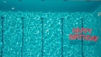 Auf dem Promotion-Foto der Vorstellung ist das wellige, leuchtend blaue Wasser eines Swimming-pools zu sehen. Unten rechts treibt ein aufblasbarer Schriftzug mit den Worten 'Happy Birthday'.