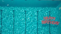 Auf dem Promotion-Foto der Vorstellung ist das wellige, leuchtend blaue Wasser eines Swimmingpools zu sehen. Unten rechts treibt ein aufblasbarer Schriftzug mit den Worten 'Happy Birthday'.