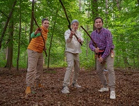 In einem grünen Laubwald stehen die drei Darstellerinnen und Darsteller der Vorstellung "bewaffnet" mit einem Stock. Sie blicken mit unterschiedlichen Mienen den Betrachtenden an.