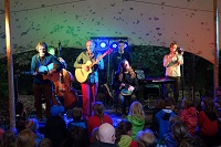 Eine Band, bestehend aus sechs Musikern (Gitarre, Chello, Posaune, Percussion, Gesang), stehen auf einer Bühne vor Publikum und geben ein Konzert.