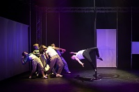 Vier Performerinnen und Performer beugen sich stark nach hinten zurück, da ein Performer, mit den Beinen an eine Metallstange geklemmt, seinen Oberkörper in Richtung der Gruppe schwingt.