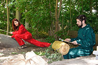 Eine Frau in rotem Overall stützt sich auf einen Baumstamm, ein Mann sitzt auf dem Waldboden und  schlägt auf eine große Trommel.