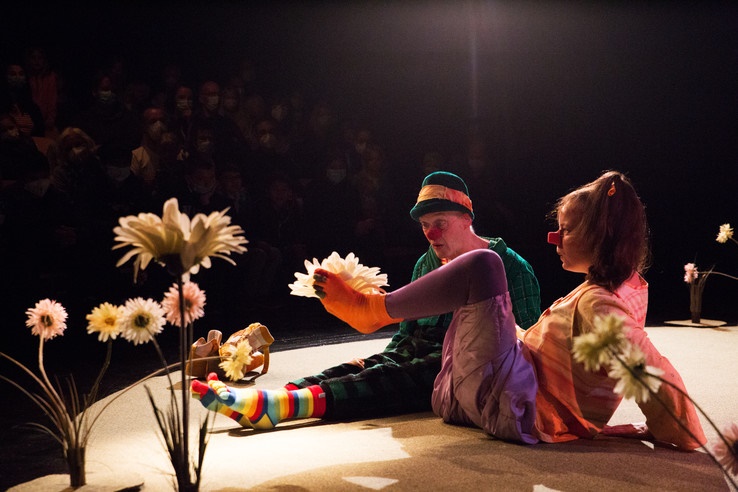Ein Clown und eine Clownin sitzen auf dem Boden. Die Clownin hält eine große Blume zwischen ihren Zehen.