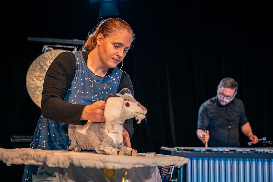 Im Vordergrund steht eine Schauspielerin, die eine Handpuppe in Form einer Ziege  hält. Im Hintergrund spielt ein Musiker auf einem Vibraphon.