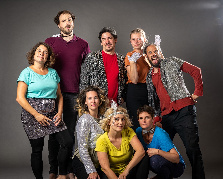 Acht festlich gekleidete Performer*innen des Ensembles 7Wiesen schauen fröhlich in die Kamera.