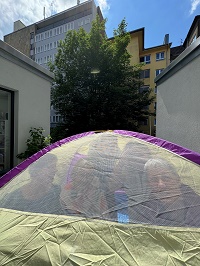 Vier Personen sitzen in einem kleinen Kuppelzelt, das in einem Innenhof aufgebaut steht. Sie schauen durch das Mückennetz in die Kamera.