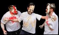 Die drei Darsteller der Inszenierung sind auf auf den Armen, dem Kopf und im Gesicht mit roter und beiger Farbe bekleckert. Sie tragen weiße T-Shirts, die ebenfalls in diesen Farbtönen beschmiert sind.