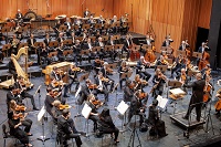 Ein Dirigent erhebt den Taktstock und leitet das 5. Sinfonieorchester Münster an. Zu sehen sind musizierende Streicherinnen und Streicher, Bläserinnen und Bläser sowie eine Harfeninstrument und ein Piano.
