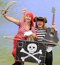 Die Musikerin, links im Bild, trägt ein Schwert und hält eine Piratenfahne. Der Musiker, rechts daneben, hält eine schwarze Gitarre in der einen Hand und ballt die Faust der anderen Hand. Beide lachen selbstbewusst in die Kamera. AHOI!