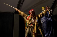 Ein Clown steht mit weit geöffneten Armen im Vordergrund und hält in der linken Hand eine Geige, in der rechten einen Geigenbogen. Hinter ihm ist eine Schauspielerin zu sehen, die ins Publikum blickt.