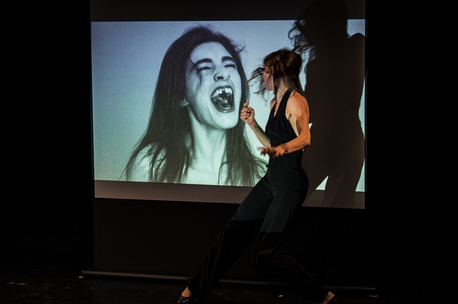 Die Performerin Cornelia Kupferschmid schaut auf eine Leinwand. Dort ist eine Frau mit schreiendem offenen Mund abgebildet.
