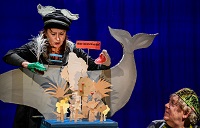 Die Schauspielerin hält in einer Hand eine Krokodilpuppe und in der anderen eine Haifischpuppe. Sie agiert vor einem Bühnenbild aus Pappfiguren.