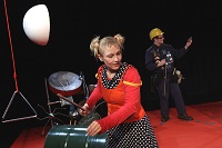 Links vorne eine Schauspielerin an einem metallenen Xylophon, hinten rechts steht ein weiterer Spieler im Hintergrund.
