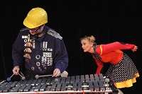 Im Vordergrund ist links vorne ein Schauspieler und Musiker an einem metallenen Xylophon zu sehen. Von hinten rechts schleicht sich neugierig die leicht gebückt Schauspielerin heran.