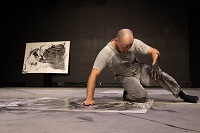 Der Schauspieler ist in grau gekleidet und kniet mit einem Bein auf dem Boden. Mit der rechten Hand stützt er sich ab. Auf dem Boden liegt ein großes Papierformat, das mit grauer Farbe mit der Hand angemalt wurde.