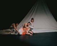 Eine in weiß gekleidete Tänzerin mit orangenem Tuch liegt auf dem Boden. Hinter ihr hocken drei weitere in weiß gekleidete Tänzerinnen und halten sie an Knien und Schultern.