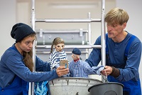 Eine Schauspielerin und ein Schauspieler, beide in blau gekleidet, halten jeweils eine Puppe in der einen Hand. In der anderen hält der Schauspieler einen Eimer, die Schauspielerin ein Handy.