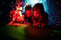 Zwei Kinder schauen sich in einer gebauten Holzwabe Miniaturinsekten an. Die Szene ist in rotes Licht eingetaucht.