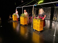 Drei ältere Frauen stehen auf der Bühne. Vor ihnen sind jeweils gelbe Kisten aufeinander getürmt, in denen sich Gemüse befindet.