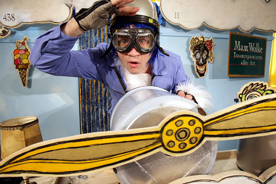 Der Spieler Christoph Bäumer sitzt hinter einem Propeller aus Pappe. Er trägt einen Fliegerhelm und eine Fliegerbrille.