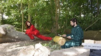 Ein in einem grünen Overlall gekleideter Musiker sitzt auf dem Waldboden und spielt auf einer Schlitztrommel. Eine in einem roten Overall gekleidete Tänzerin macht einen langen Ausfallschritt auf einen Baum und schaut zum Musiker.