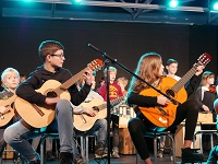 Gitarrenschülerinnen und - schüler sitzen im Profil auf einer Bühne und spielen ihr Instrument.