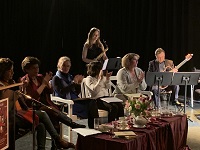 Mehrere Personen sitzen um einen Tisch auf einer Bühne, im Hintergrund sind drei Musiker/innen zu sehen.