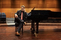 Foto: Jürgen Bleibel sitzt mit überschlagenen Beinen auf einem Klavierhocker, den Arm auf einem schwarzen Flügel aufgestützt.