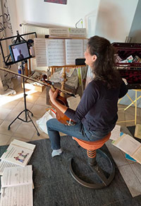 Eine Frau mit Geige sitzt vor einem Notenständer, auf dem ein Tablet liegt, in dem eine Geigenschülerin zu sehen ist.
