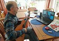Mann mit Gitarre sitzt an einem Tisch, auf dem ein Notebook steht. Darin ist eine Schülerin bei Gitarrenspiel zu sehen.