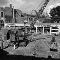 Schwarz-weiß-Foto eines Baggers auf einer Baustelle vor einer eingeschössigen Ladenzeile