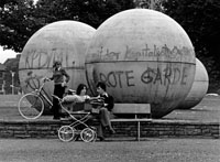 Vor drei großen Betonkugeln sitzt ein junges Paar mit Kinderwagen auf einer Bank und steht eine junge Frau mit Fahrrad