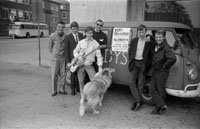 Sechs junge Männer, von denen einer eine Gitarre hält, stehen mit einem Collie vor einem VW-Bus