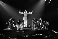 Auf einer Theaterbühne knieen mehrere Schauspielerinnen um den Hauptdarsteller des Musicals Jesus Christ Superstar herum, der in seinem weißen Anzug mit weit ausgebreiteten Armen in Segnungsgeste wie ein leuchtendes Kreuz erscheint.