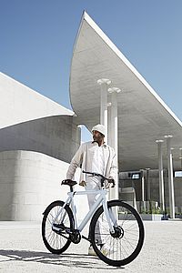 Fotografie eines Mannes in weißer Kleidung mit einem weißen Fahrrad mit schwarzen Reifen vor futuristischer Architektur