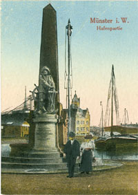 Auf einer colorierten Fotopostkarte steht ein Paar in historischer Kleidung vor dem Hafendenkmal