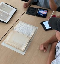 Schüler sitzen um einen Tisch, auf dem Archivalien ausliegen. Daneben liegen auch Tablets der Schüler.