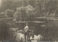 Schwarz-weiß-Foto von 1918: Ein Mann und zwei Kinder stehen an einem großen Teich, im Hintergrund ist ein Floß mit fünf Männern darauf zu sehen.
