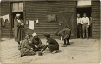 Kriegsgefangene vor einer Baracke aus Holz hockend und in der geöffneten Tür stehend.