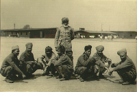 Foto von Kriegsgefangenen, eine Gruppe Männer mit dunkler Haut und Turbanen hockt am Boden, hinter ihnen steht ein etwas älterer Mann mit Turban und weißem Bart.