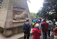 Gruppe von Personen an einem Denkmal aus Stein, in dessen Sockel ein liegender Löwe gemeißelt ist, der Schriftzug darüber 'Treue um Treue' ist mit Farbe beschmiert.