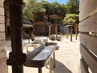 Zwischen zwei Holzbauten steht ein Wasserspiel mit Röhren und Wannen.