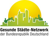 Logo des Gesunde Städte-Netzwerk der Bundesrepublik Deutschland Gelbe Sonne, die über einer in Grün gezeichneten Stadt aufgeht