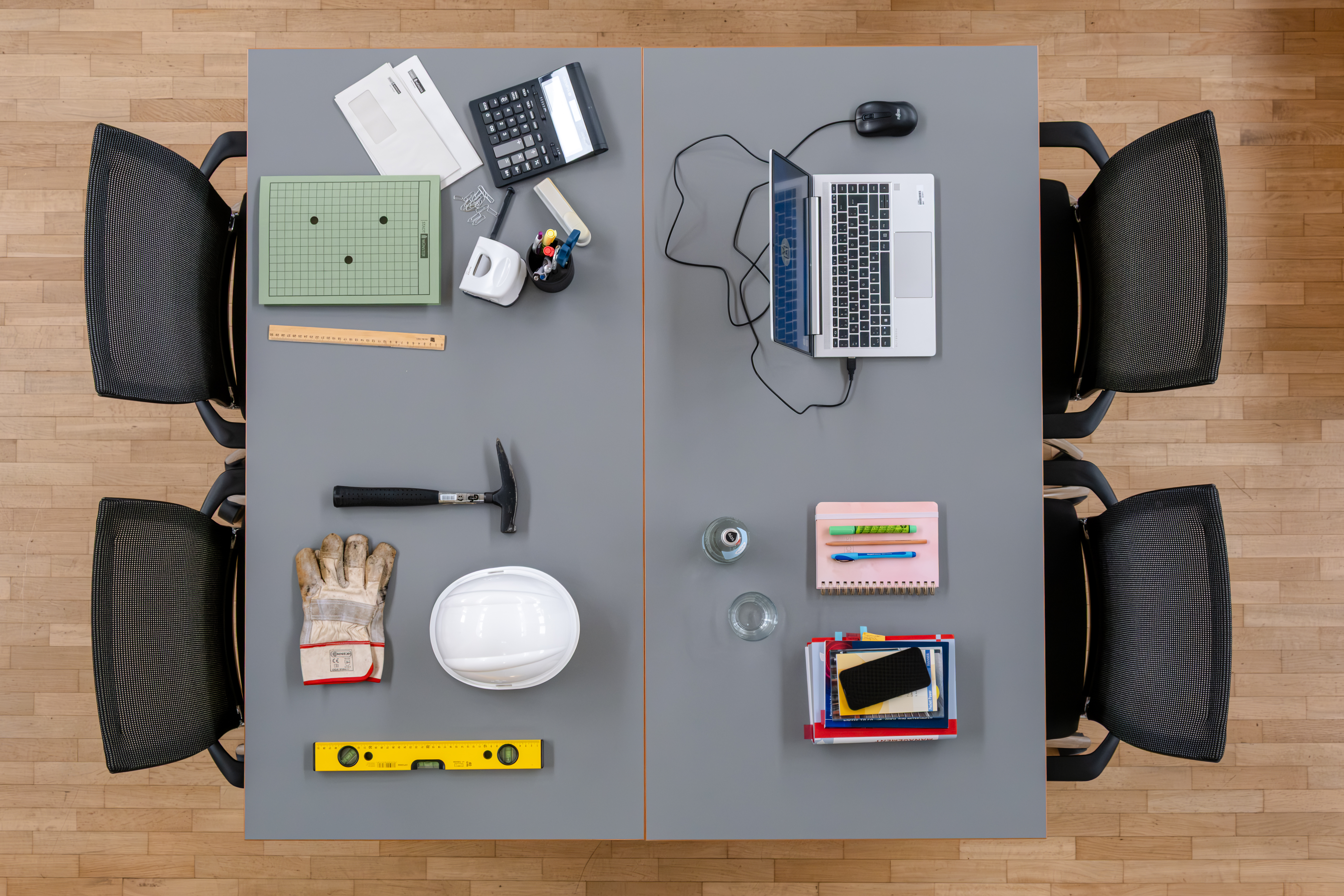 Ein Tisch mit vier Arbeitsplätzen von oben fotografiert: Auf dem Tisch liegen verschiedene Arbeitsutensilien, wie ein Hammer, Wasserwaage, aber auch Laptop und Handy.