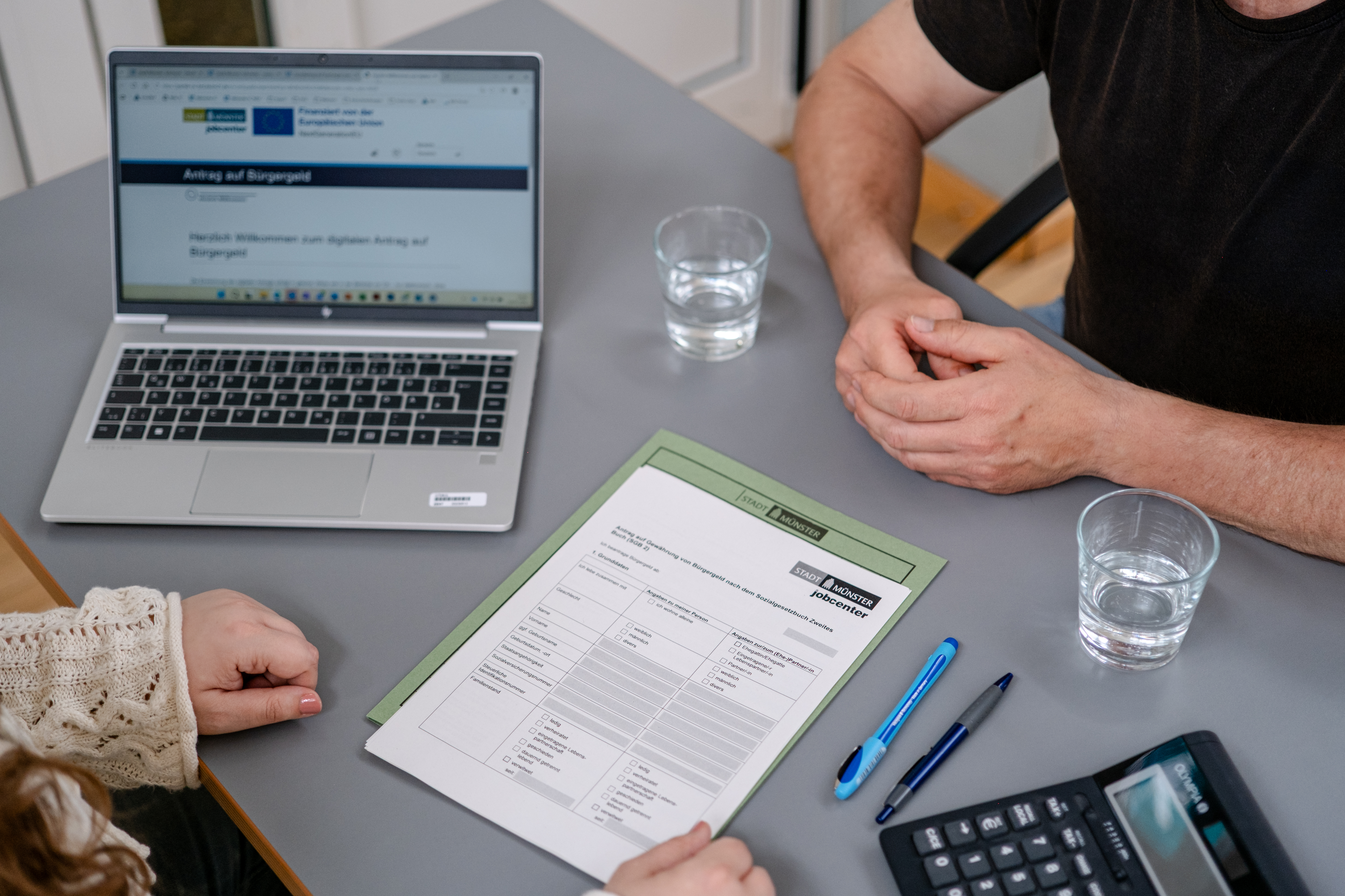 Zwei Personen sitzen an einem Tisch sich gegenüber. Vor ihnen liegt ein Papier, daneben steht ein Laptop und ein Taschenrechner.