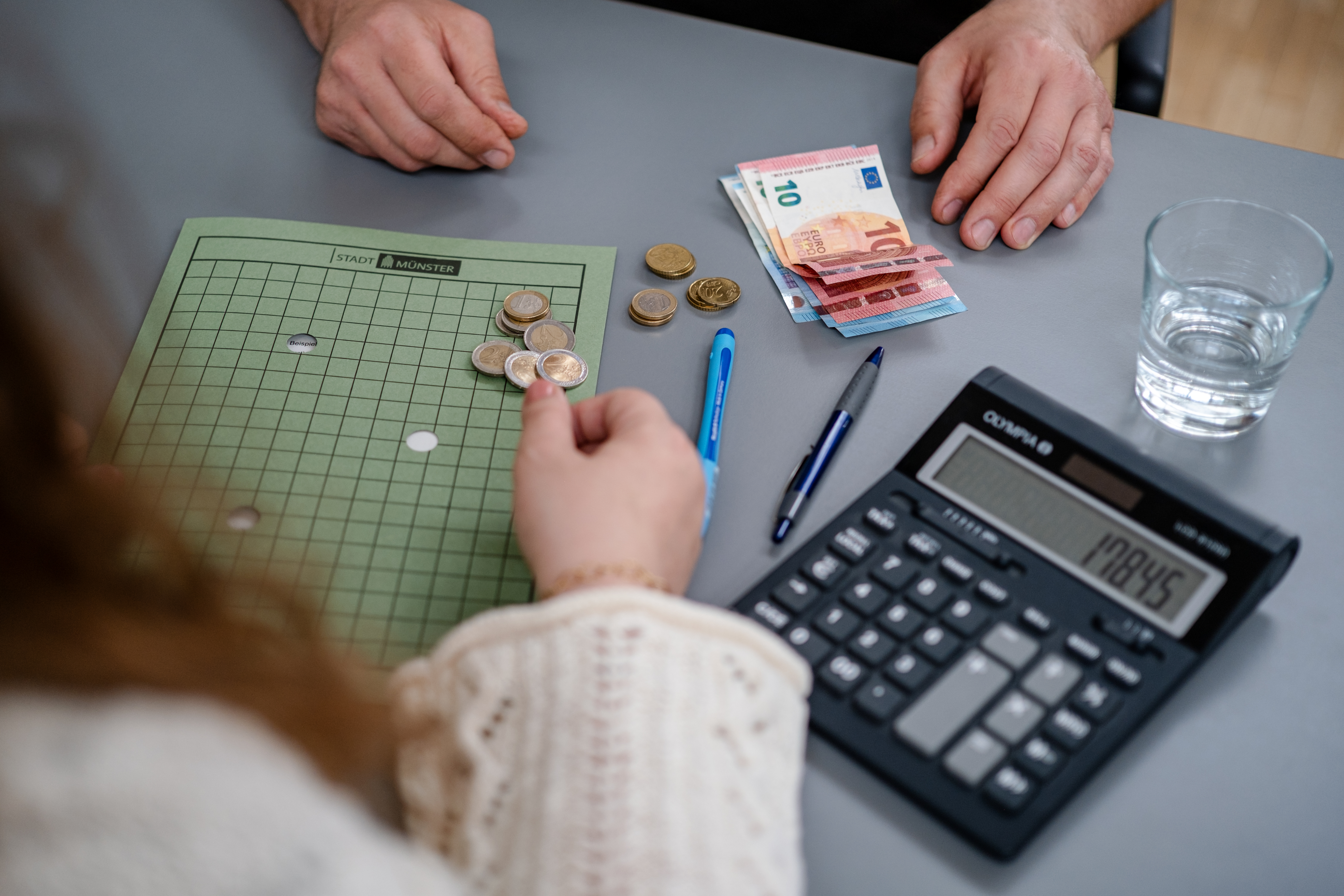 Zwei Personen sitzen an einem Tisch. Auf dem Tisch liegen ein Taschenrechner, eine Arbeitsmappe und Geld.