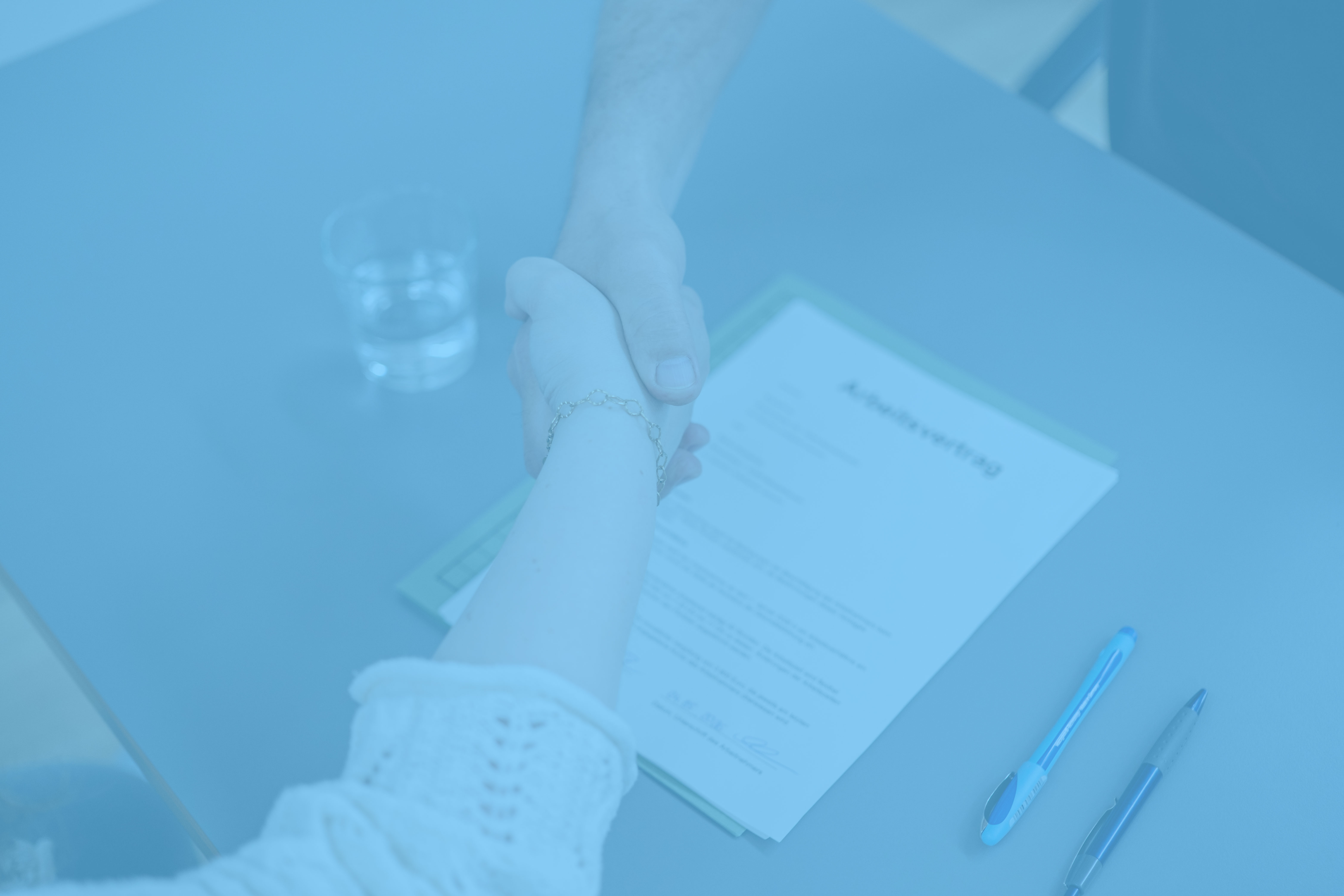 Nahaufnahme eines Handschlags von zwei Personen. Ein Arbeitsvertrag liegt darunter auf einem Tisch. 