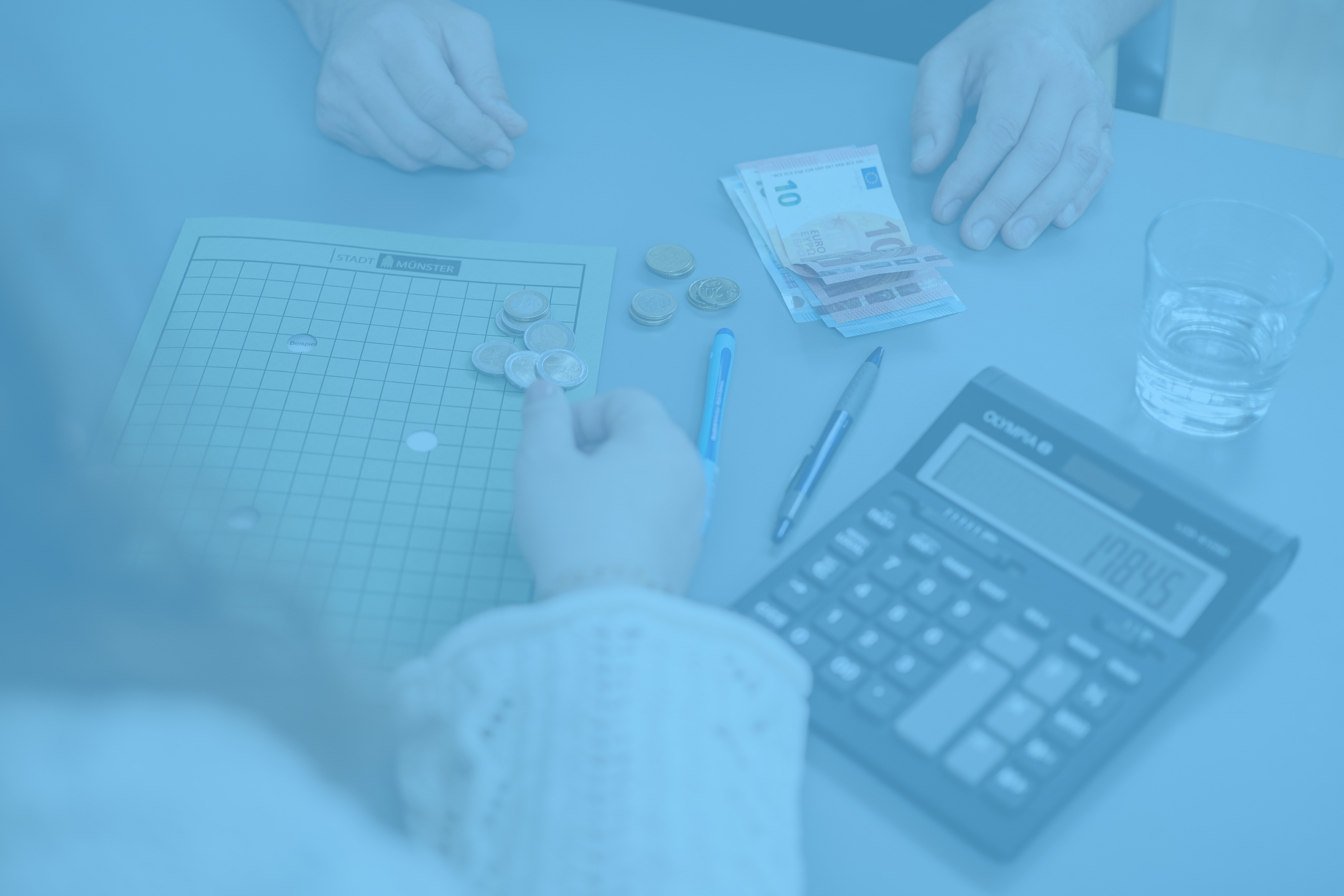 Zwei Personen sitzen an einem Tisch. Auf dem Tisch liegen ein Taschenrechner, eine Arbeitsmappe und Geld.