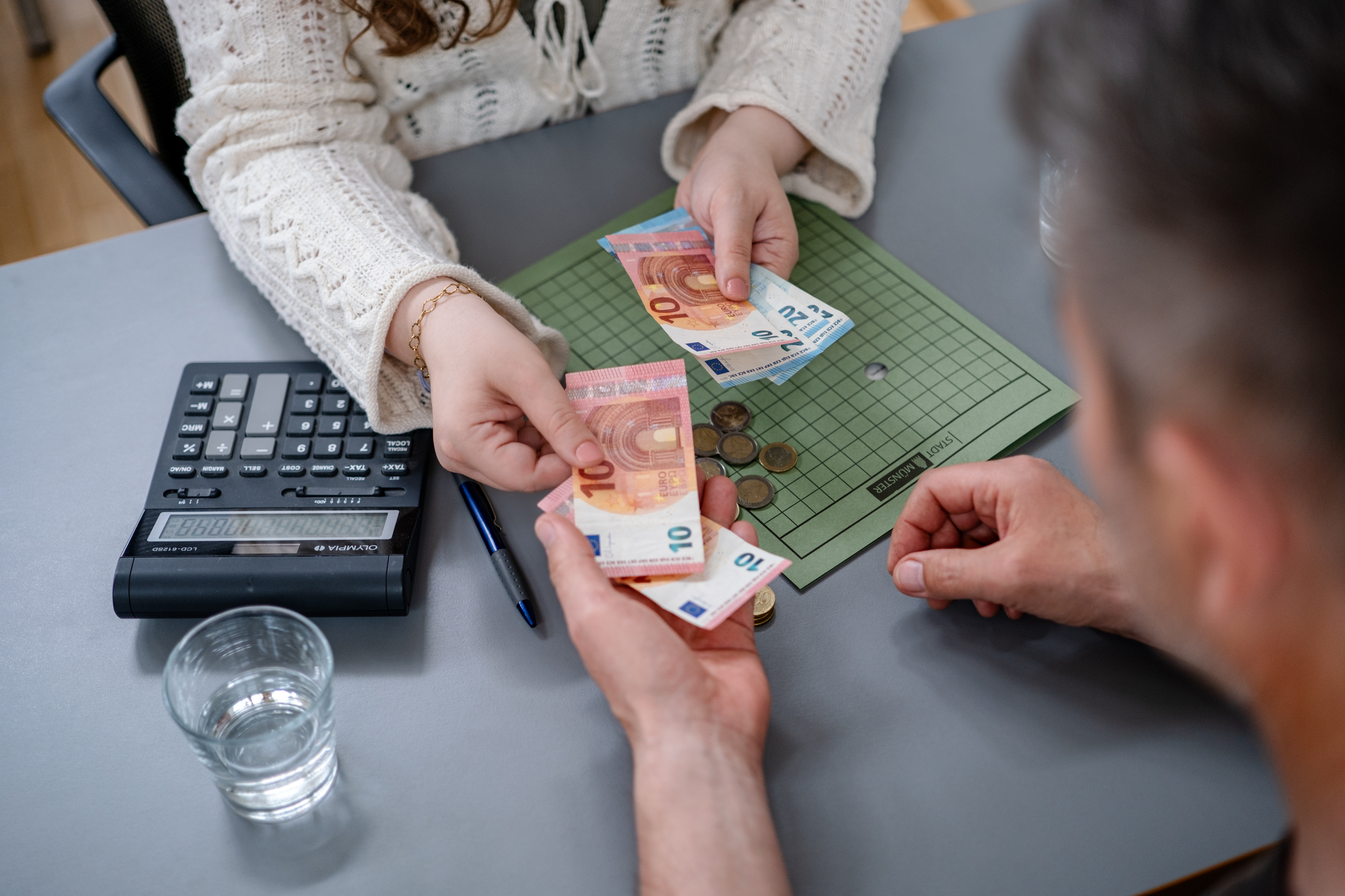 Zwei Personen sitzen an einem Tisch. Eine Hand reicht der anderen Geld.