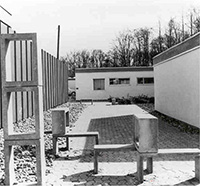 Schwarz-weiß-Foto: Gepflasterter Platz mit kubischen Betonskulpturen  vor schlichten Flachbauten